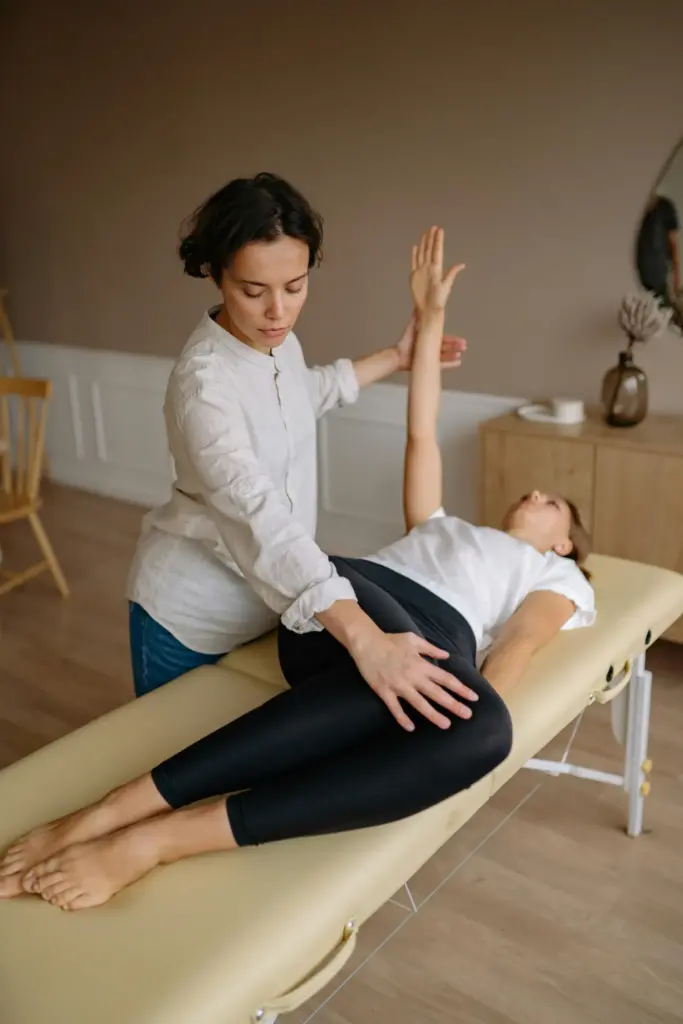 A person on a massage table at home is being guided through a stretching exercise by another individual in a room with wooden flooring and minimal decor.