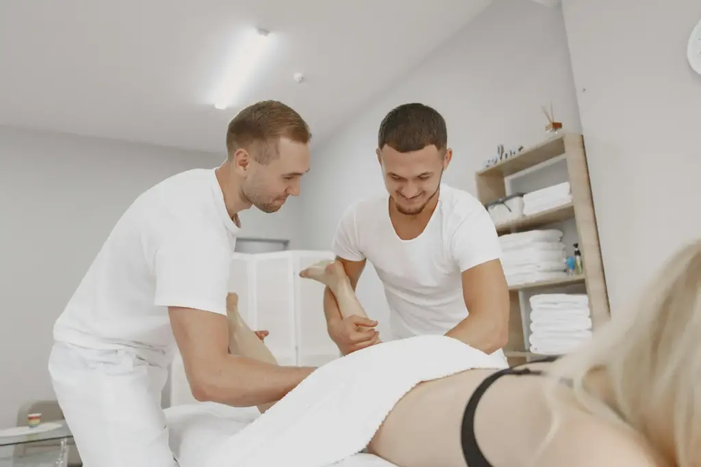Two massage therapists work together on a client lying face down, covered by a white towel in a cozy home treatment room with shelves of towels in the background.