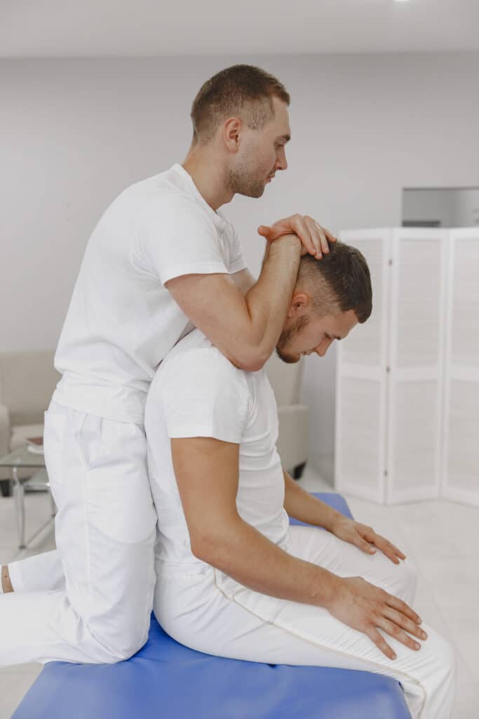 In a clinical setting, a man in a white outfit kneels on a table, performing a therapeutic neck stretch on another seated man, both dressed in white, emphasizing the importance of long-term care.
