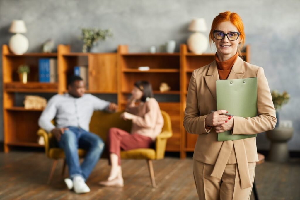 A woman with red hair holding a clipboard stands in the foreground. In the background, a couple sits on chairs in front of wooden shelves.