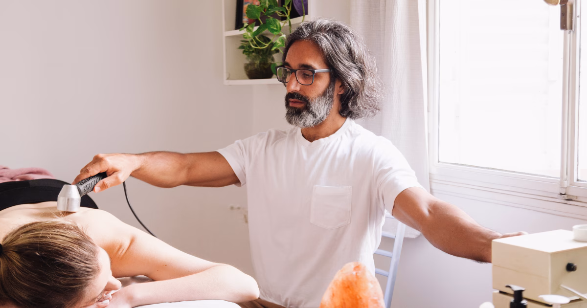 A therapist uses an ultrasound therapy device on a patient's back in a well-lit treatment room.