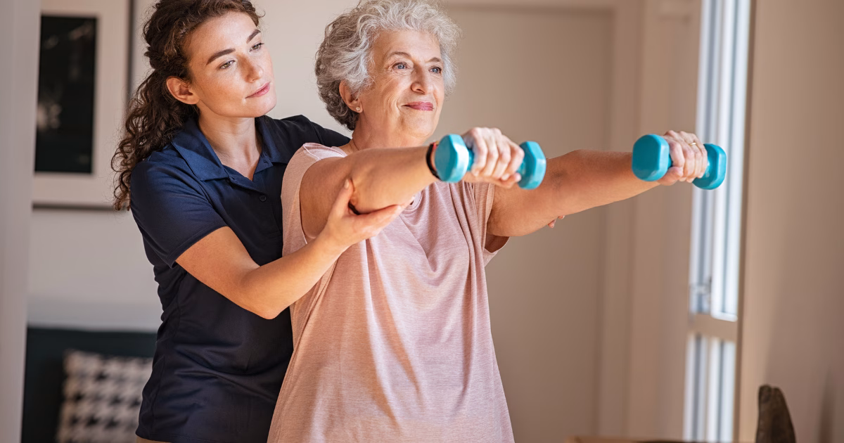 A younger woman assists an older woman in lifting light blue dumbbells, helping her exercise in a well-lit room.
