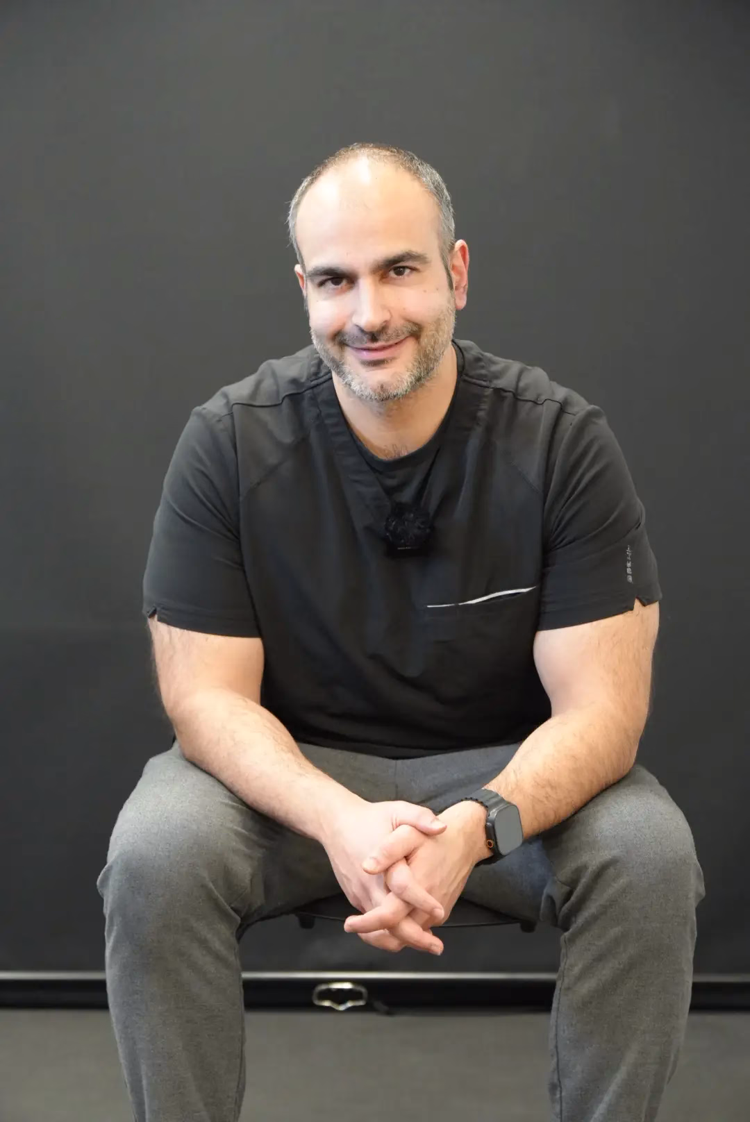 A man with short hair and a beard, wearing a black shirt and gray pants, sits on a stool against a plain dark background, smiling with his hands clasped—reflecting the approachable spirit of Mobile Therapy.