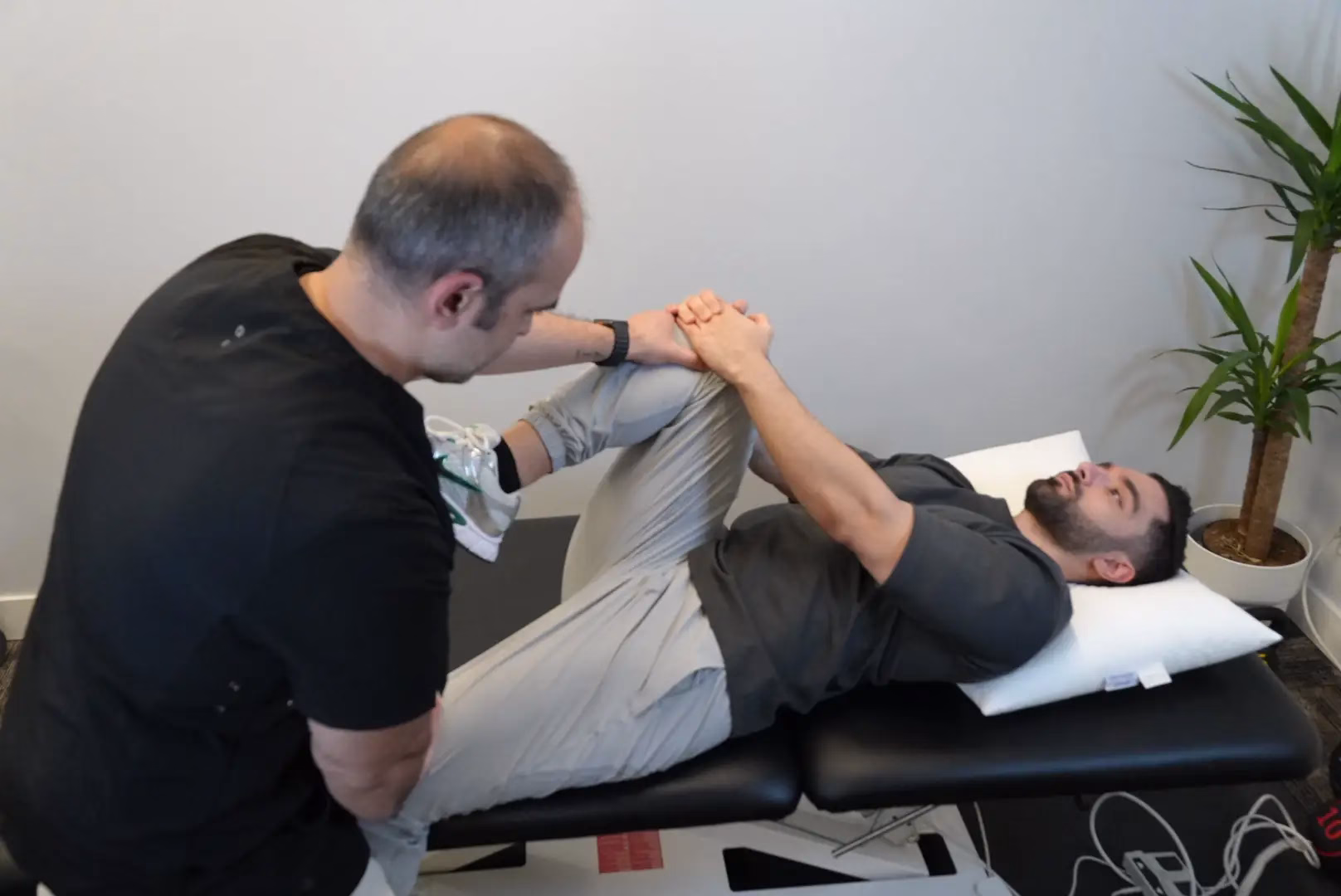 A physical therapist from a Mobile Physiotherapy service assists a man lying on an exam table with a knee and hip stretching exercise in a clinical setting.