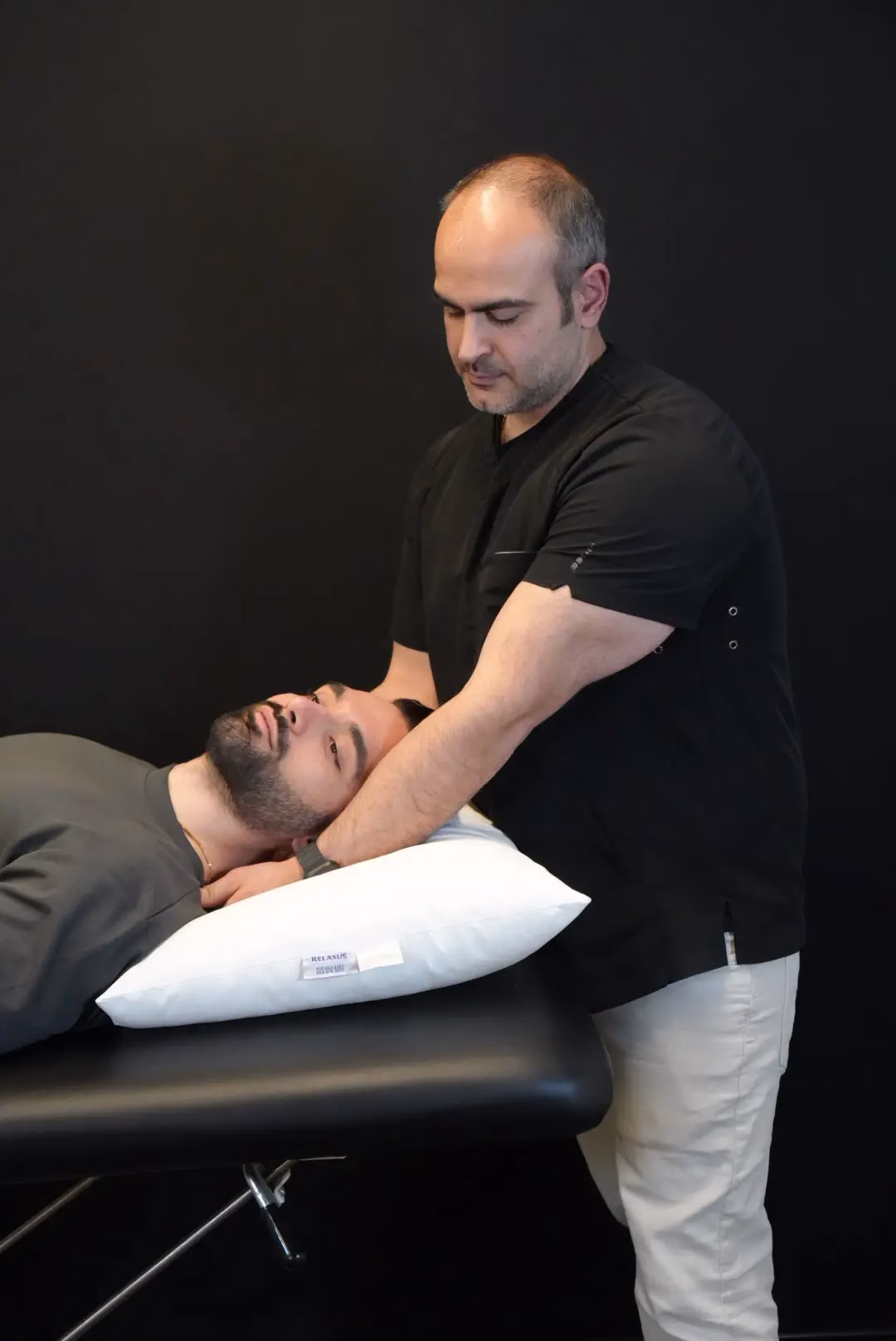 A man lies on a table with his head on a pillow while another man, standing, performs a neck adjustment or examination as part of kinesiology services.