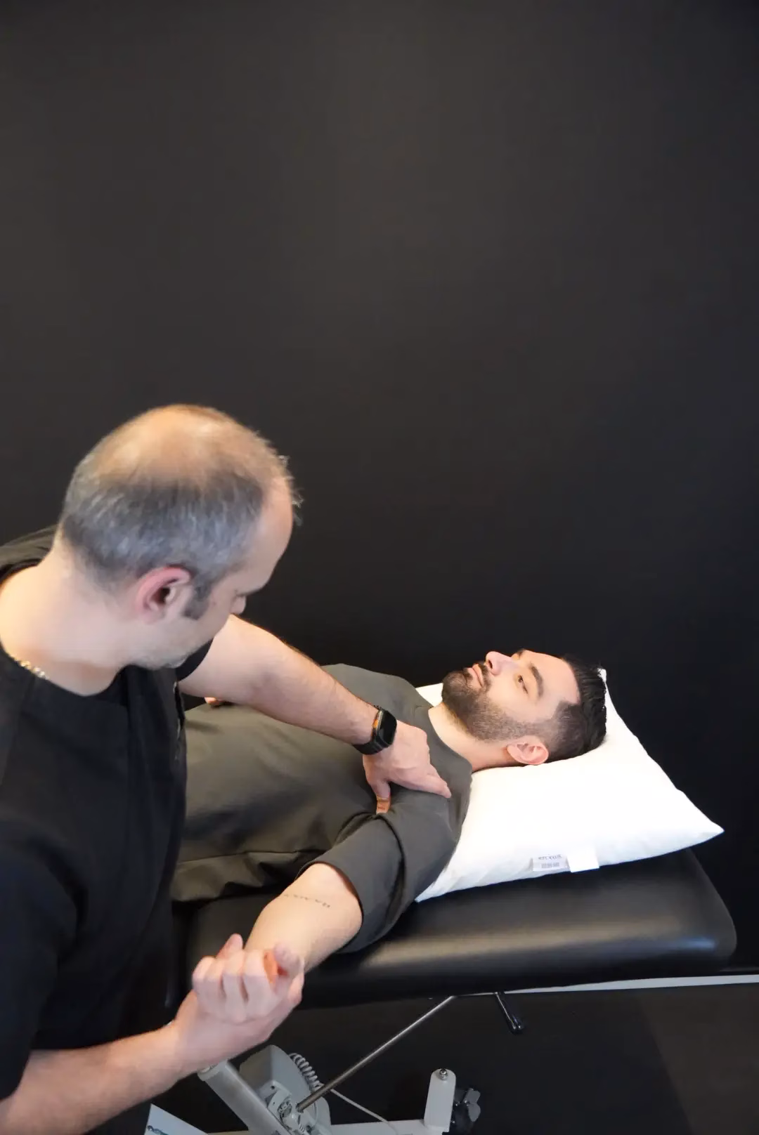 A man lies on an exam table while another person in black presses on his upper chest, performing a physical examination of the shoulder area as part of kinesiology services.
