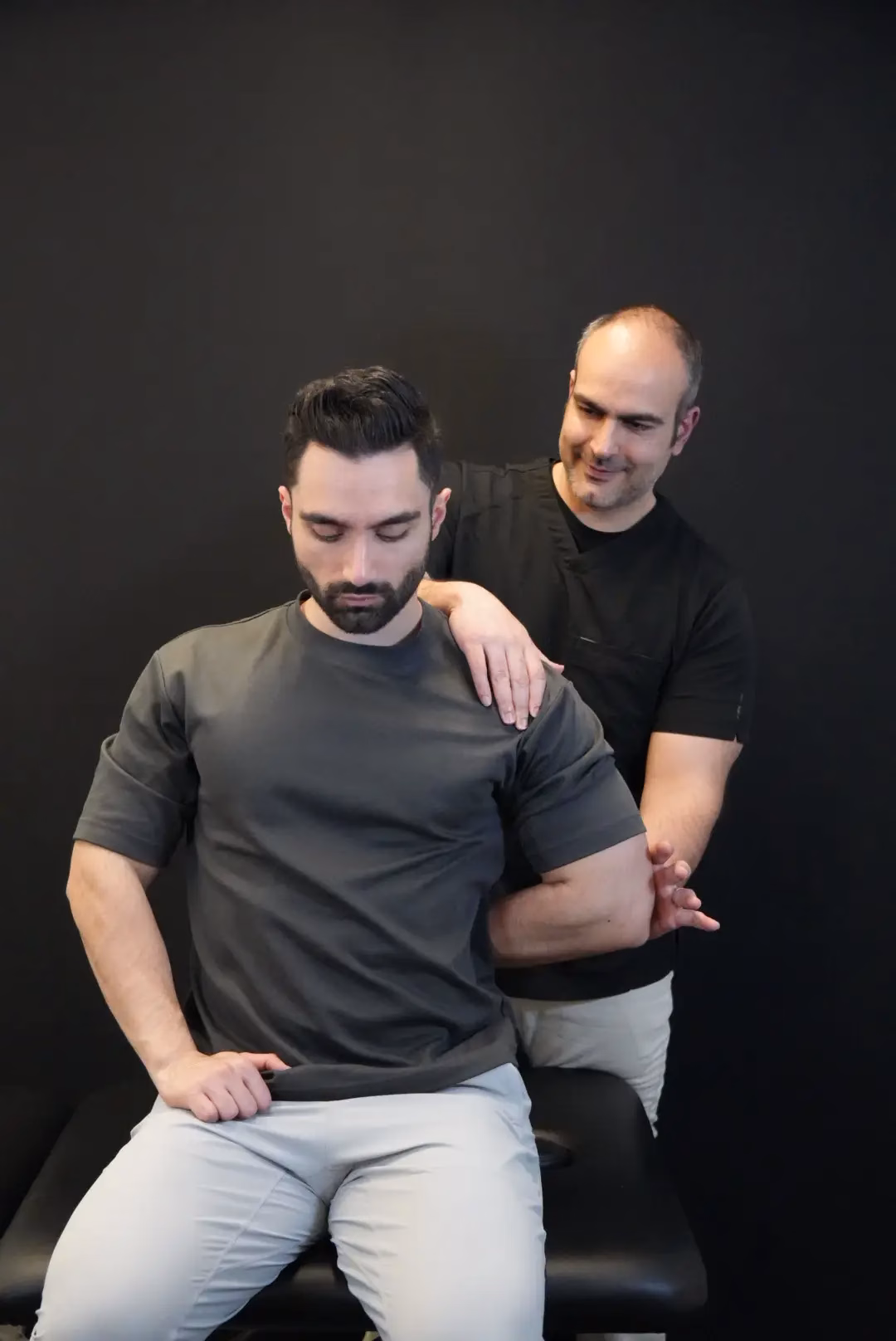 A man performs a shoulder stretch with assistance from another man against a plain black background, showcasing the benefits of Mobile Therapy Services.