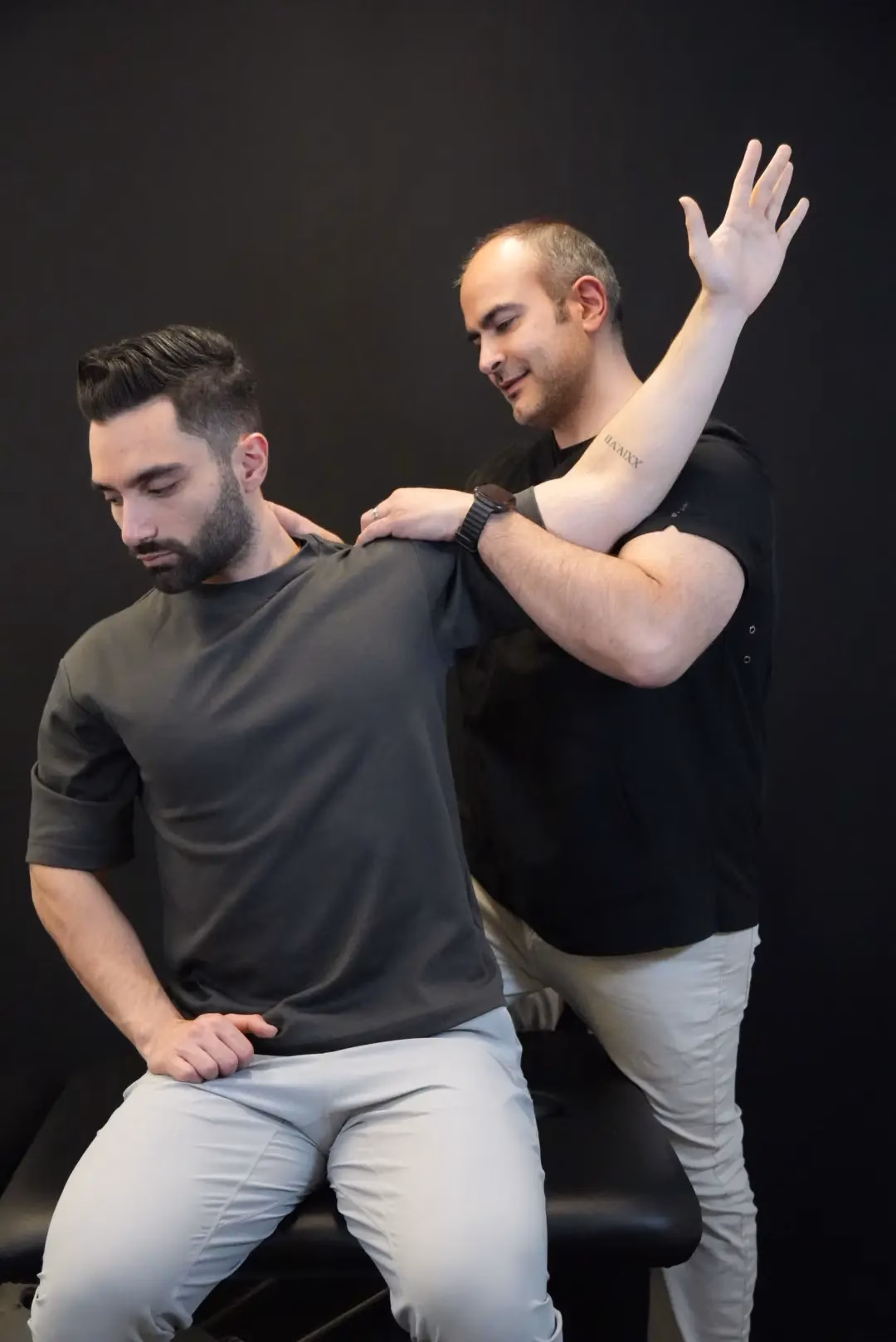 A physical therapist from Mobile Therapy Services assists a seated man by lifting and stretching his arm against a black background.
