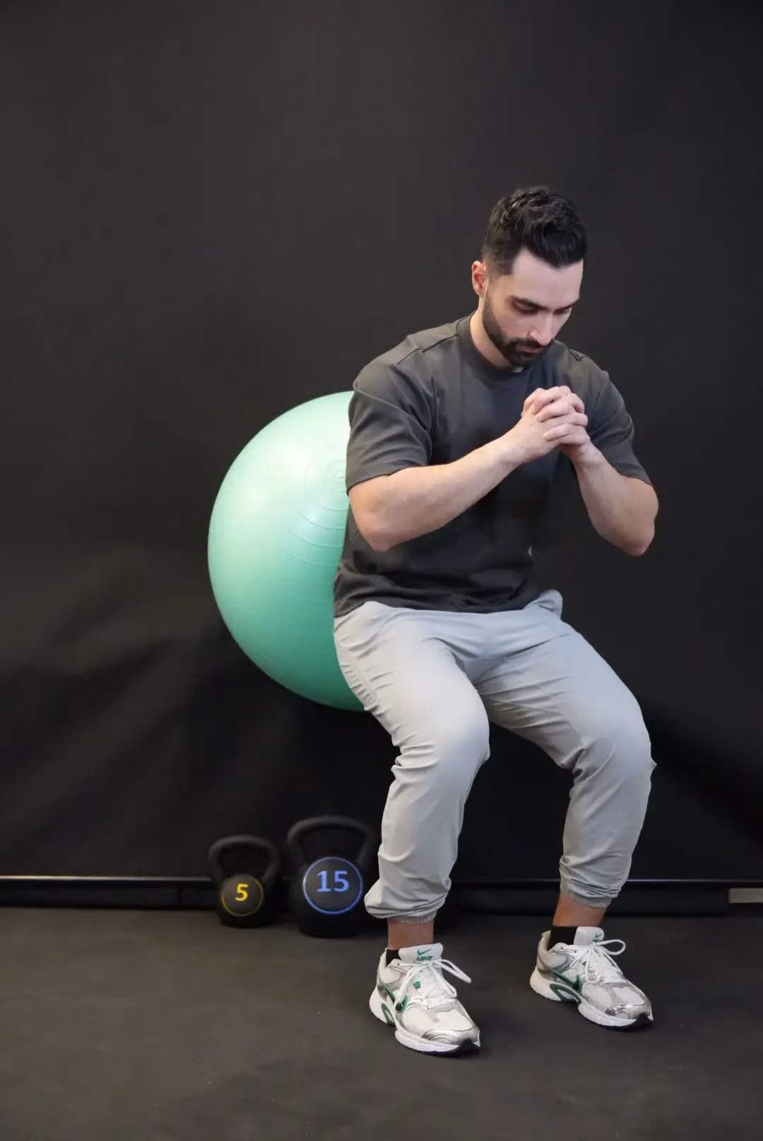 A man performs a wall squat with a stability ball behind his back, hands clasped, and two kettlebells nearby—an exercise often recommended by Mobile Physiotherapy experts in Vancouver.