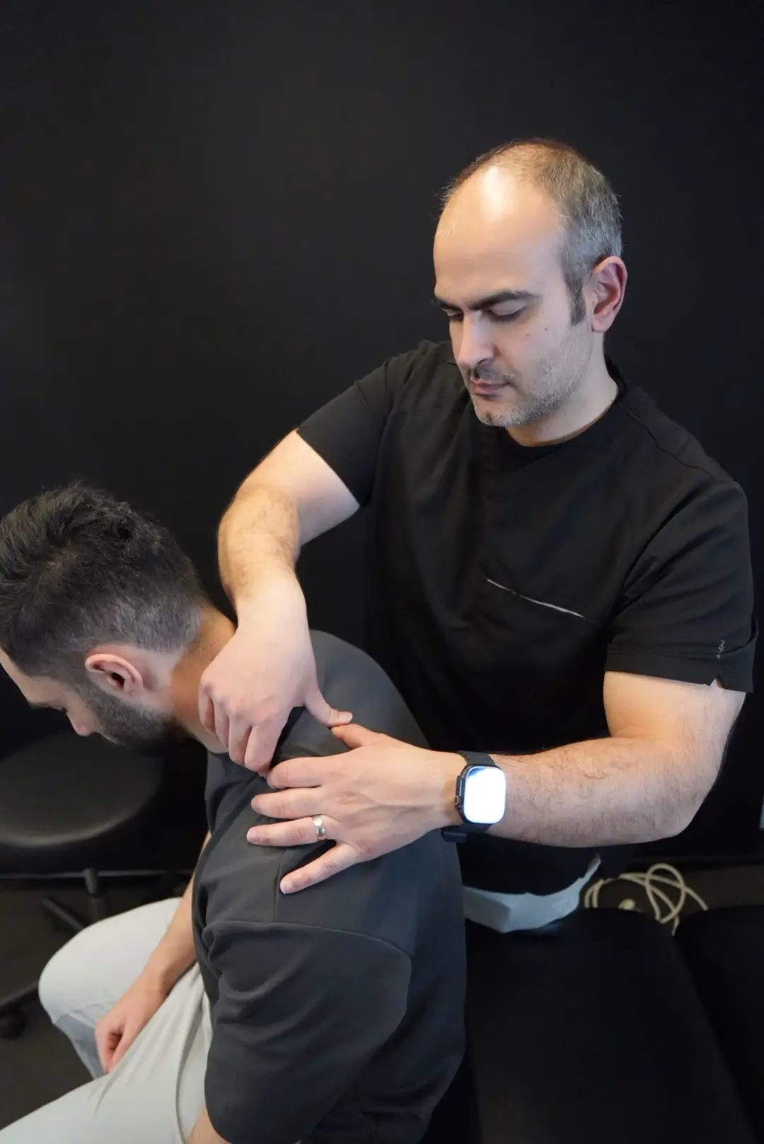 A person in black scrubs performs a manual neck and shoulder adjustment on a seated man in a dark shirt against a black background, showcasing expert care by Mobile Physiotherapy Vancouver.