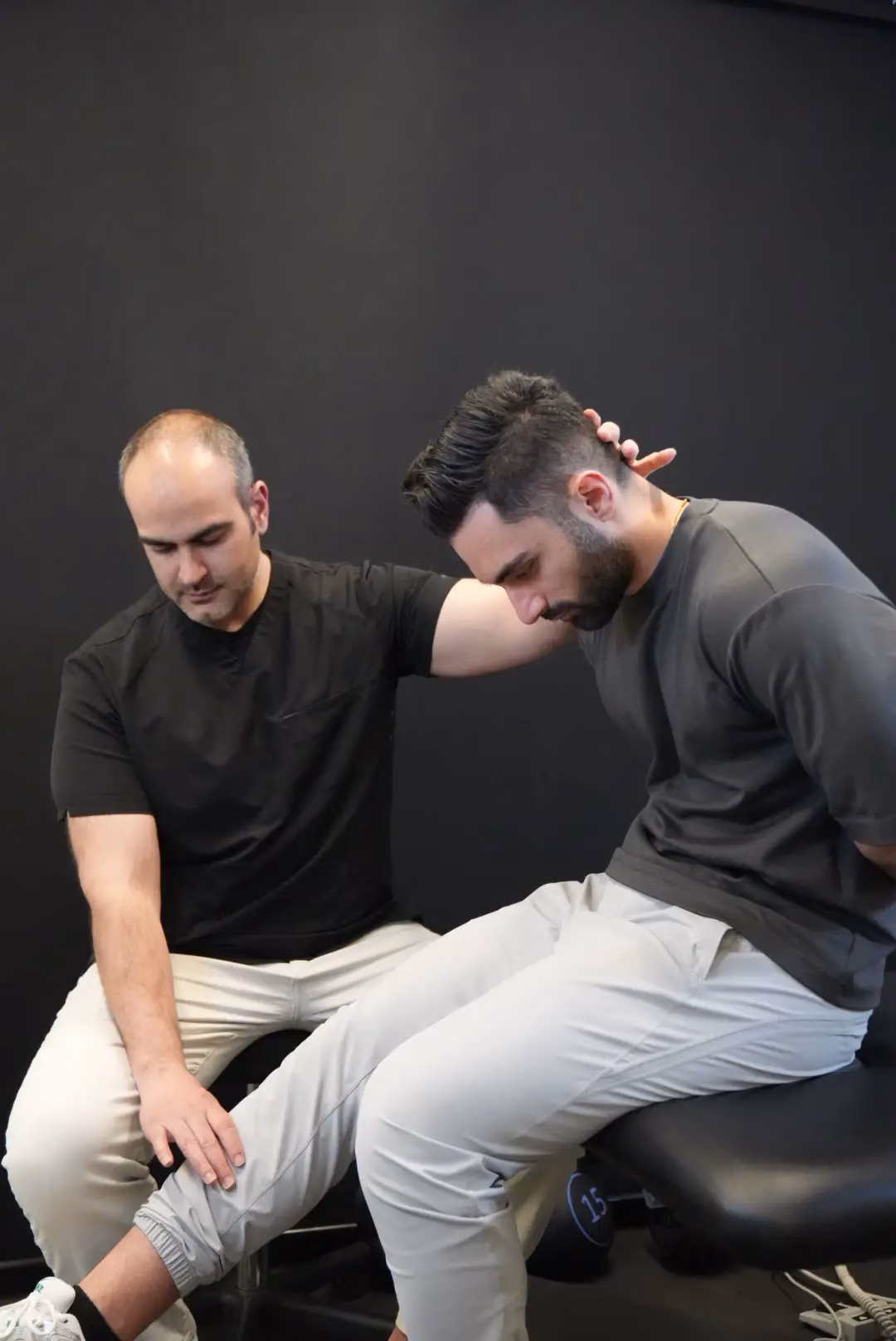 A physical therapist from a Vancouver mobile physiotherapy service performs a seated stretch on a patient, holding his neck and knee, against a plain black background.